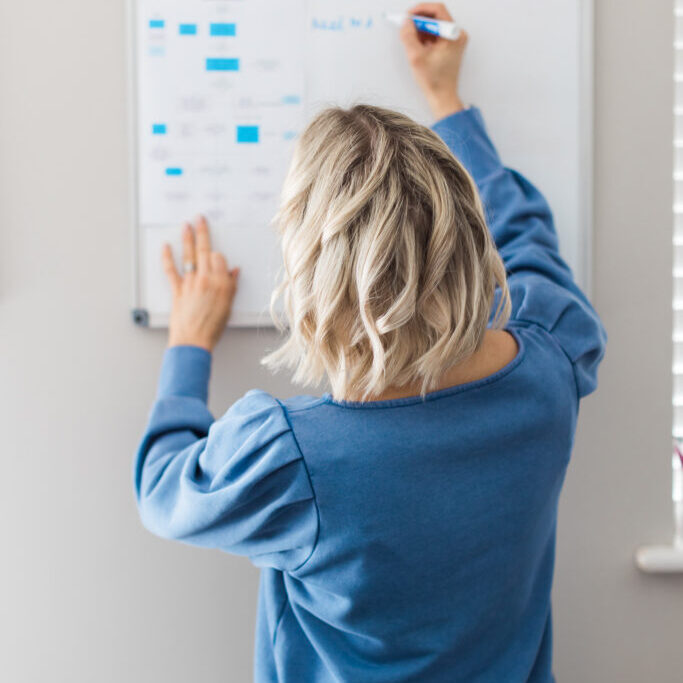 Photo of Kellie standing in blue jumper creating a process map on a whiteboard.